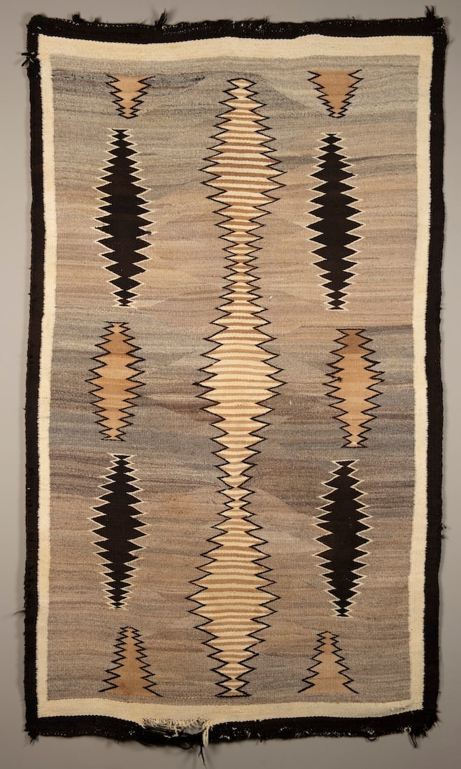 A Navajo (Diné) rug: 1930s, the black border surrounds serrated lozenge devices on either side of a larger central serrated lozenge, wool loss at the ends. 82 x 47 1/2 in.