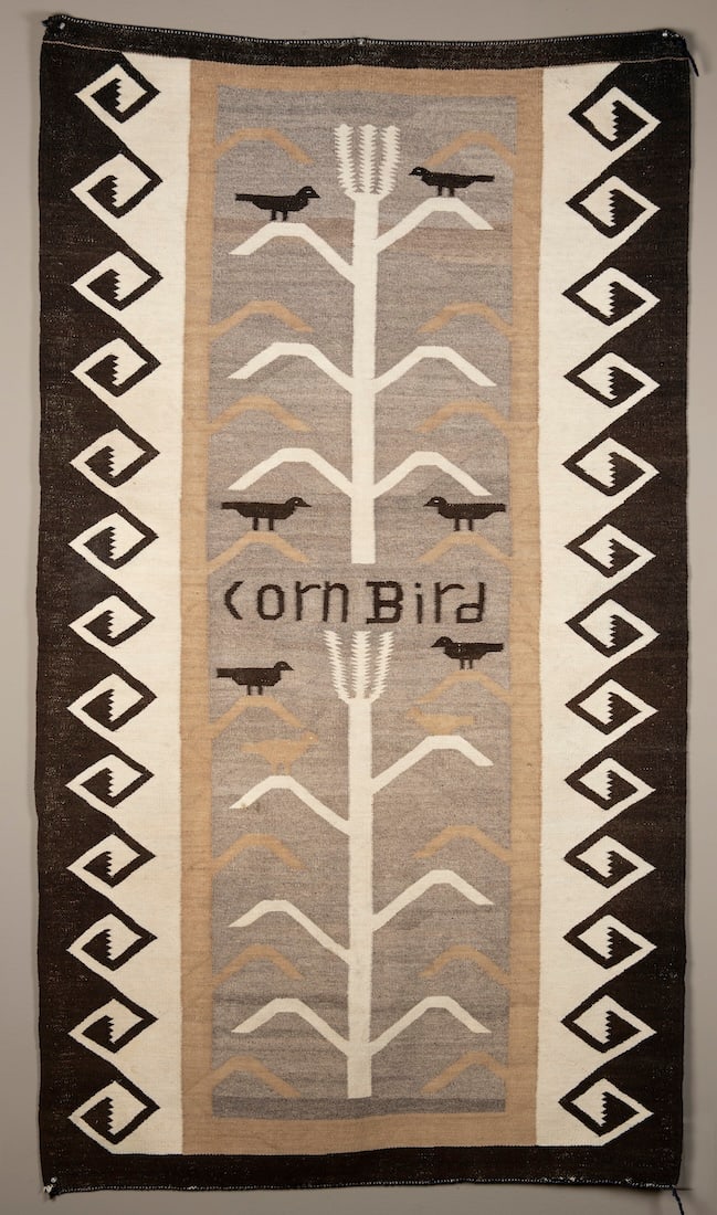 A Navajo (Diné) effigy rug: The sides with black border and diamond key design, the center with a single corn stork with six resting birds in black and “Corn Bird”, written in the center. 73 1/2 x 47 in.