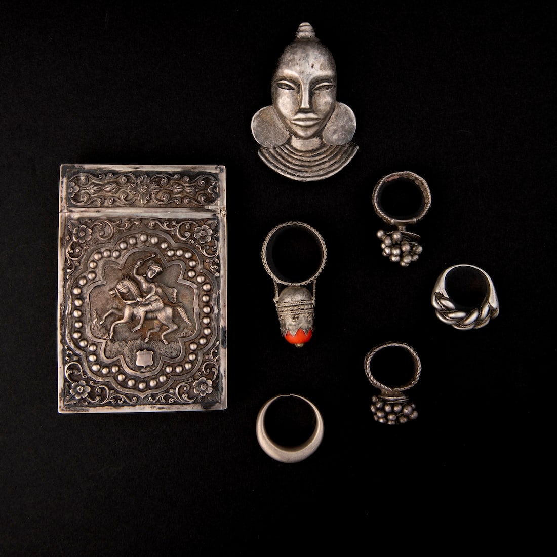 A collection of ethnic jewelry: A cigarette case with Indian motifs on each side, an African clip pendant of a woman's head, and five rings. Size of case 4 x 2 3/4, lg. of pendant 2 3/4, ring sizes 14.5, 11.5, 9.5, 9.5, and 7.5.