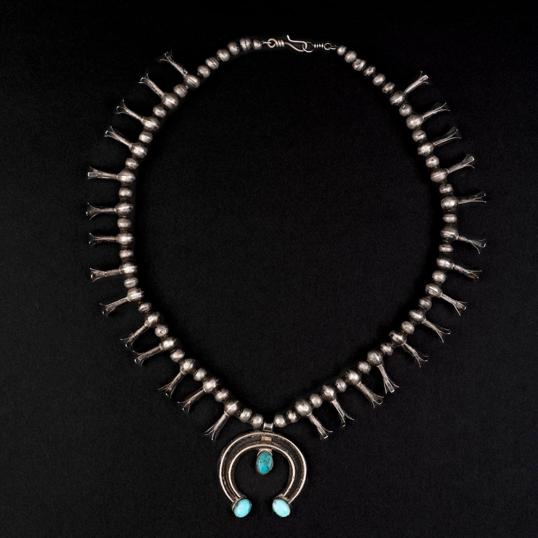 A Navajo (Diné) Silver and turquoise squash blossom necklace: Second quarter 20th century, the silver bead neckalce with twenty-eight squash pendants, the two-wire naja with three turquoise settings. Lg. of necklace 19 1/2, lg. of naja 1 3/4 in.