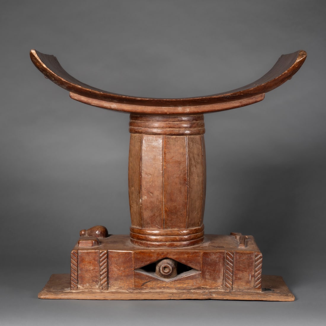A large wood Ashanti stool: Akan, Ghana, early 20th century, the large wood stool, carved from one piece of wood, the rectangular base with a rifle, powder flask and box carved in high relief, the barrel-like pedestal supports a