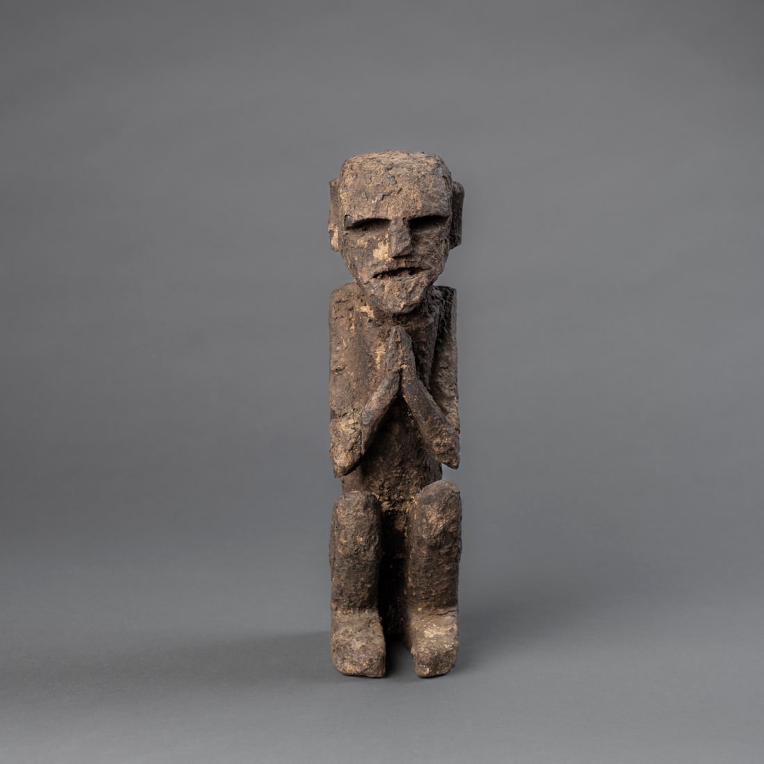 A West Nepal Shaman figure: A carved wooden figure depicting a seated man with hands clasped in prayer or invocation, the face is strongly formed, with deep-set rectangular eyes, a broad flattened nose, and open mouth. The surfa