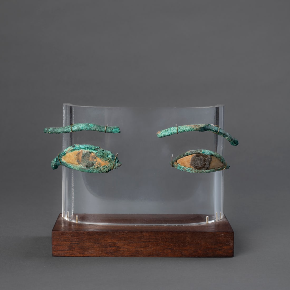 A pair of Egyptian bronze eyes from a funerary mask: New Kingdom, circa 300 B.C., A pair of cast bronze eyes, each formed with almond-shaped outline and stylized brows above. The surfaces show areas of deep green and blue patina. Mounted together on a m