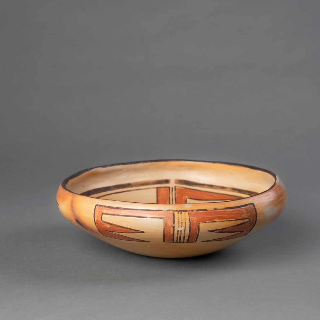A Hopi polychrome bowl: Shallow vessel with incurved rim and suspension lug, painted on the interior with a stylized bird in red and black on a tan slip, enclosed within geometric elements. Exterior decorated with black and