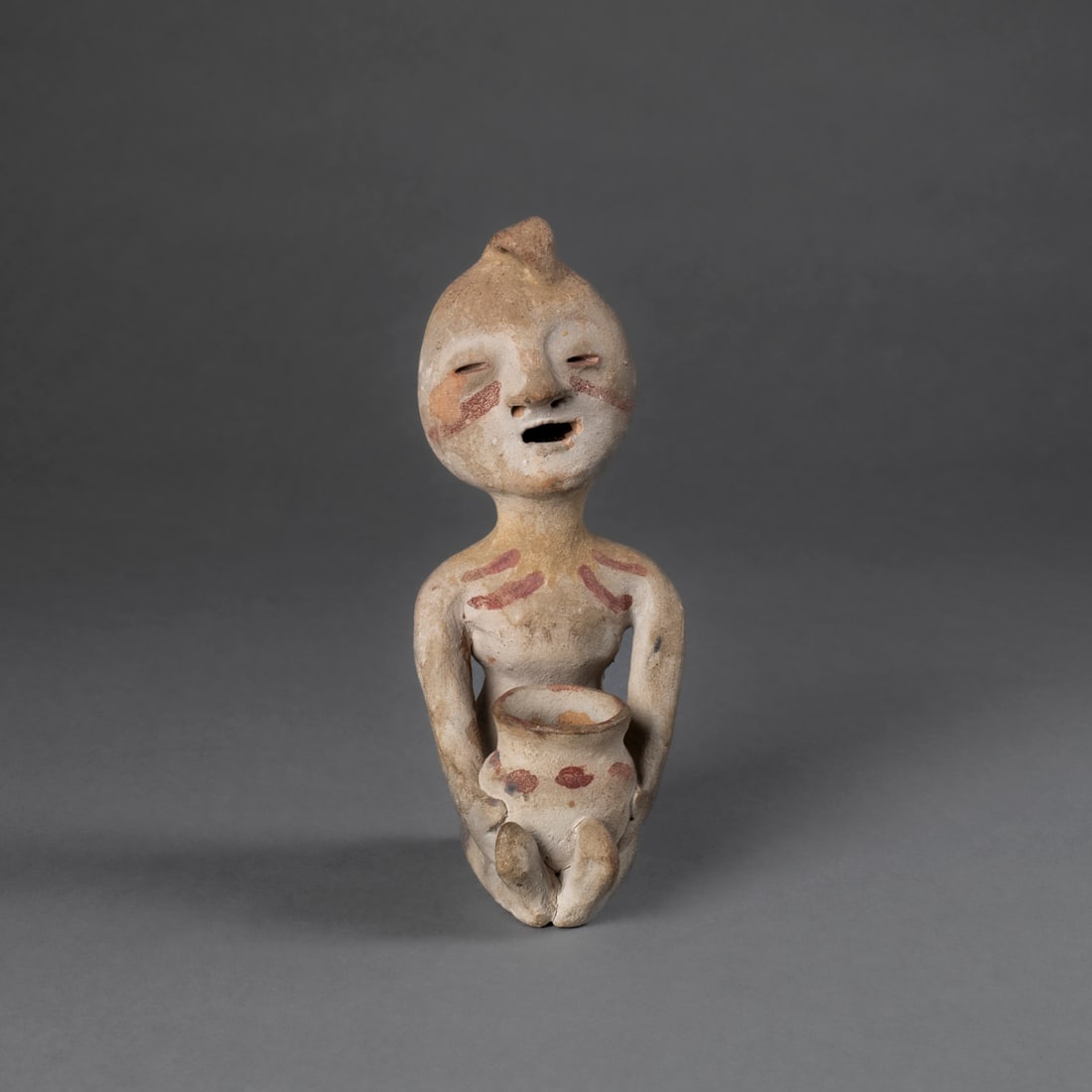 A Cochiti Rain God: The formed gray colored clay in the shape of a rain god holding a water vessel with red motifs, and red painted lines on the stomach and on the face. The facial features incised, and a suspension hand