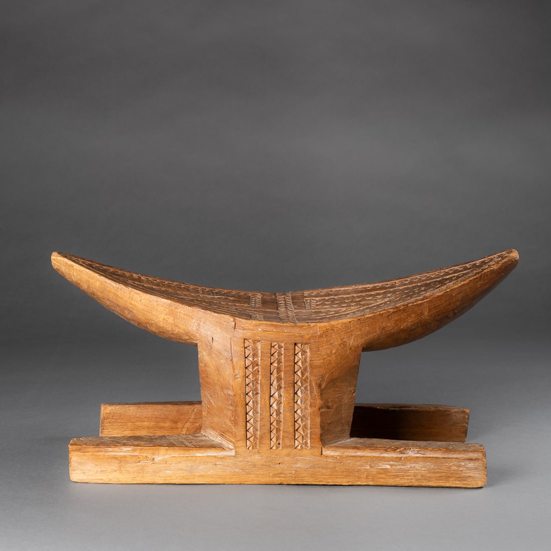 A Suriname stool: Hand-carved hardwood stool, featuring a double triangular seat supported by a central plank base. The seat and sides are decorated with incised geometric patterns in zigzag and chevron motifs. Smooth