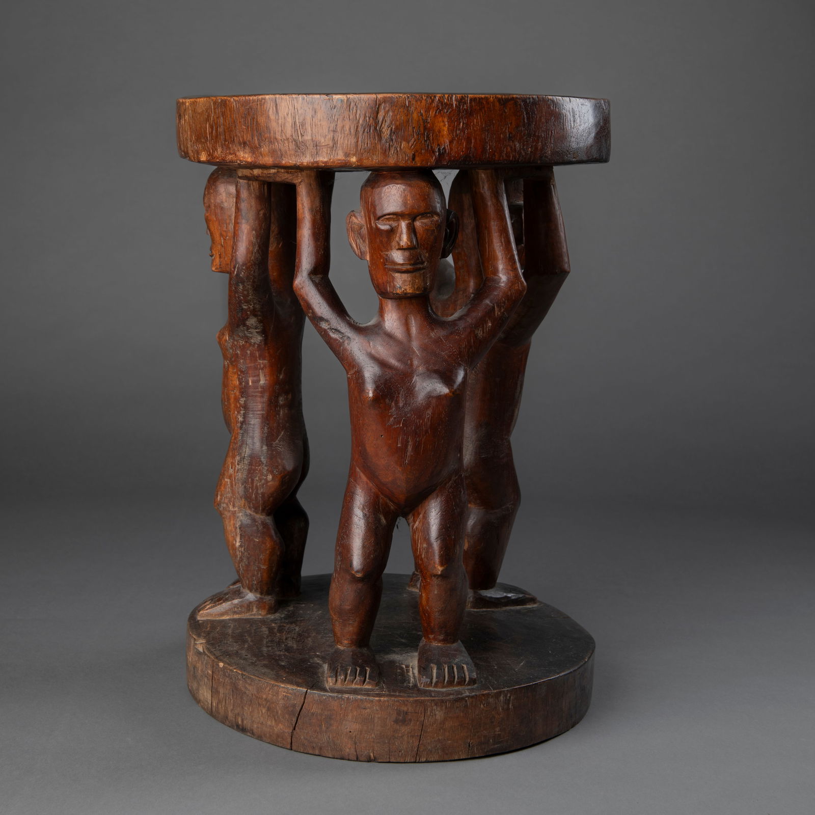 A Philippines effigy stool: Early 20th century, the heavy hardwood stool, with three standing figures supporting the heavy round seat, dark smooth patina. Ht. 19 1/2, wd. 13 1/2 in.