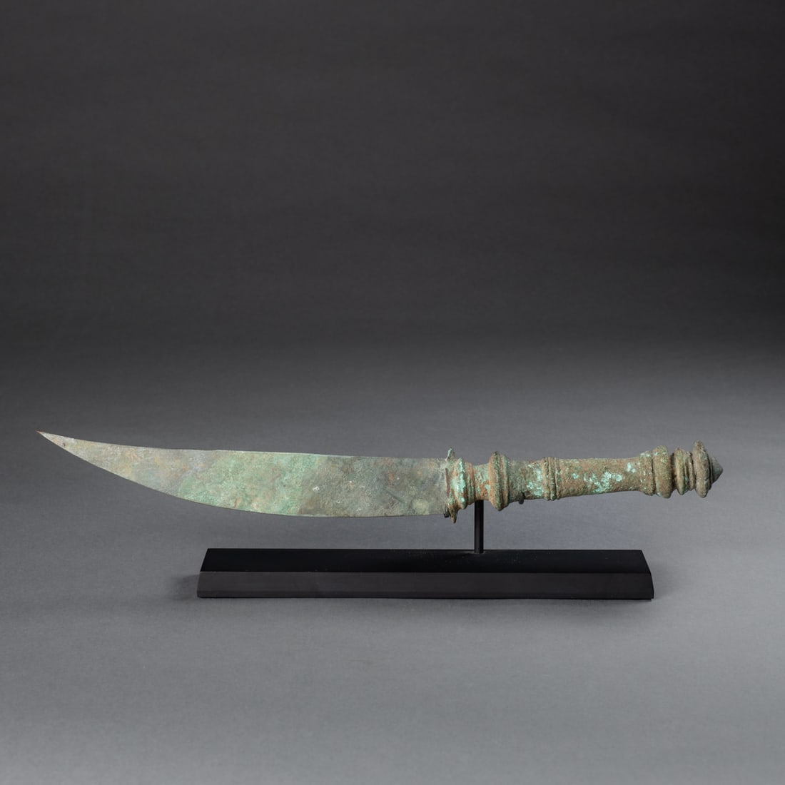 A Khmer bronze dagger: Ankor Thom, circa 11-12th century, A.D., the bronze blade with flat top side and curved blade, with round handle with round cone-shaped pommel, mounted on a metal base. Lg. 13 1/2 in. Provenance: