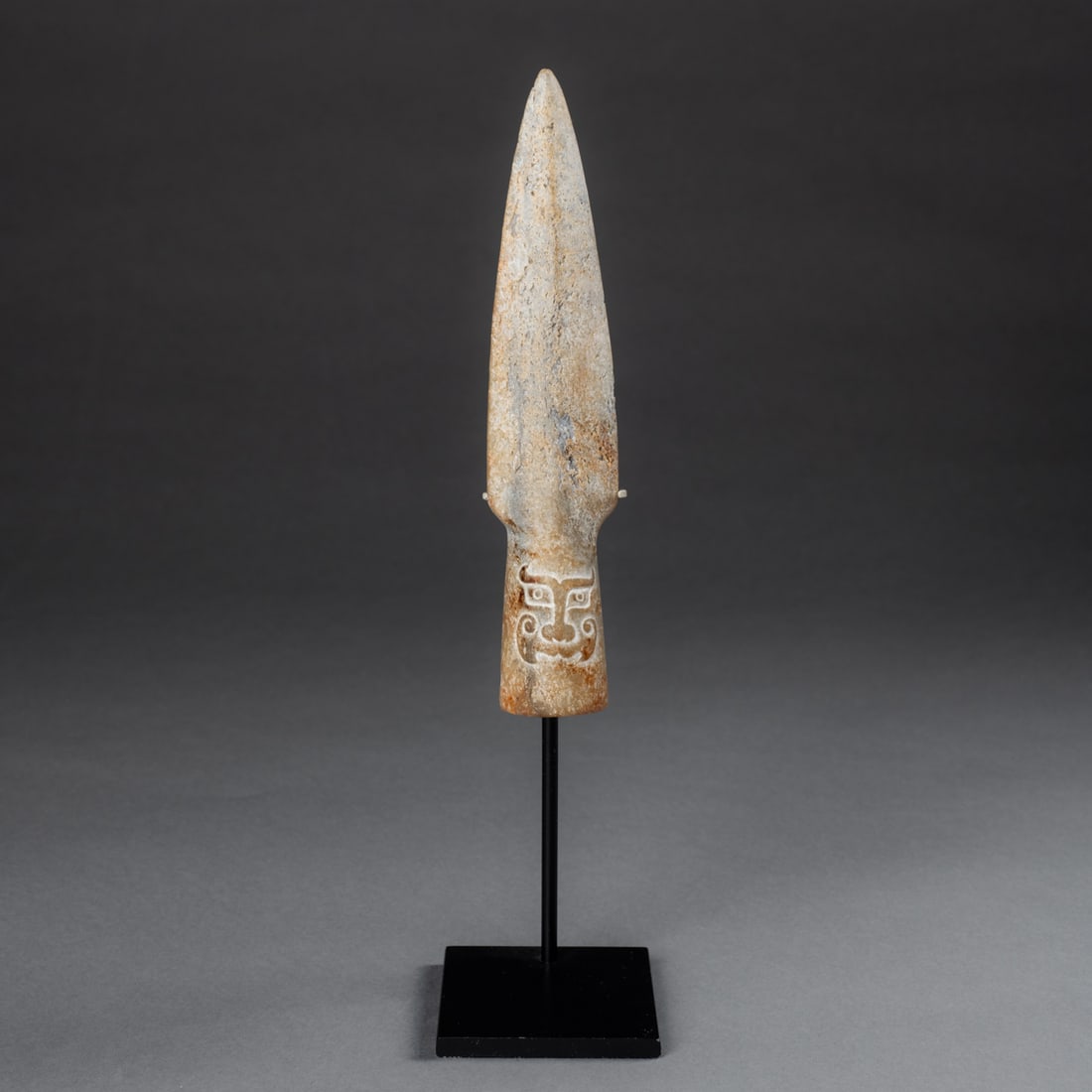 An Erlitou Neolithic Chinese spear point: Circa 1900-1500 B.C.E., a carved stone spear point, with carved face on the one side, mounted on a custom metal stand. Ht. 7 1/4, wd. 1 1/2 in. Provenance: Private collection, Albuquerque, NM; John Wh