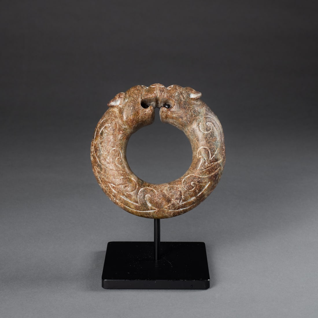A Shang Dynasty stone ring: Circa 1600-1100 B.C.E., the stone ring decorated with a dual pig and dragon motif, mounted on a custom metal stand. Dia. 5, wd. 1 3/8 in. Provenance: Private collection, Albuquerque, NM; John Whelan F
