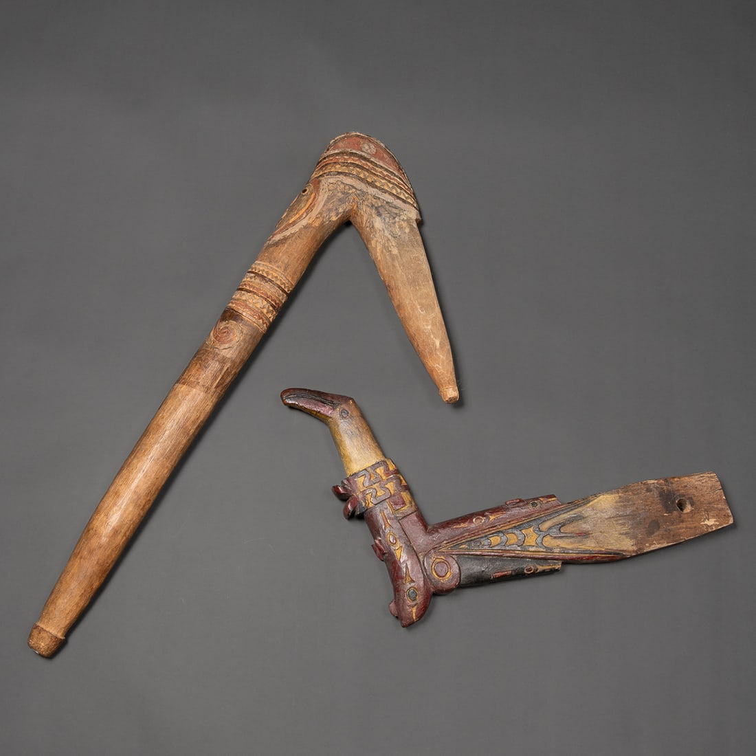 Two New Guinea items: The first carved and painted Maprik wood axe handle with anthropomorphic face at the angled head, featuring wide eyes, open mouth, and raised motifs in red and black pigments. The second carved and pa