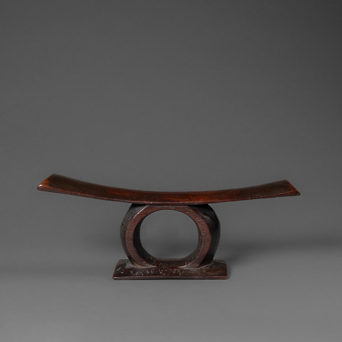 A Fijian neck rest: 19th century, standing on a low rectangular base, the gently curving pillow supported by a circular projection from the base, with old collection label on the underside, dark, smooth patina. Ht. 5 1/2