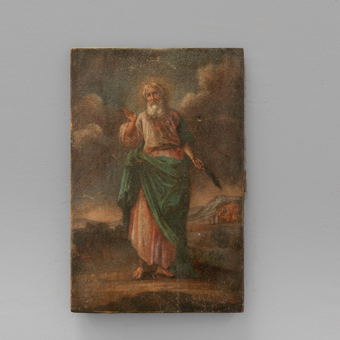 An 18th century Spanish colonial retablo: Painted on wood depicting a standing male saint with glowing halo, right hand raised in blessing or prayer, wearing a green garment under a long white robe. Set against a clouded background with