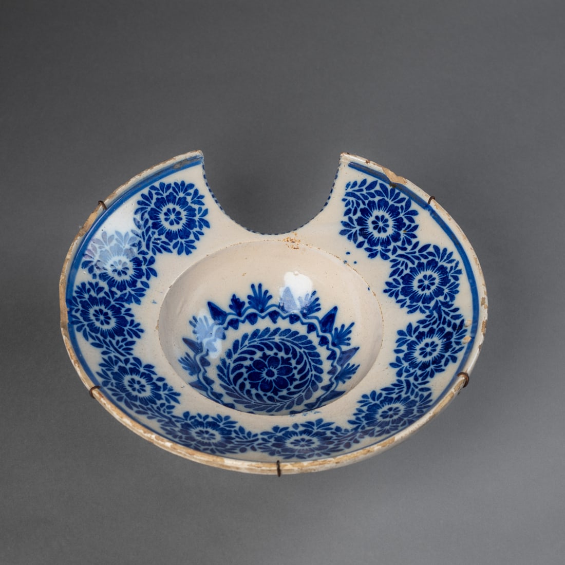 A 19th century Spanish barber's bowl: Circular earthenware bowl with crescent shaped cut-out rim, glazed in cobalt blue with a band of floral medallions circling the rim and in the center. The underside retains wire reinforcement for stab