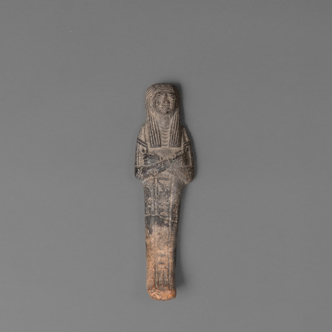 An Egyptian sculpture: Clay figure depicting a standing mummy with crossed arms at the chest and long straight headdress, the face finely detailed with incised features. Smooth wear consistent with age, lightening near the