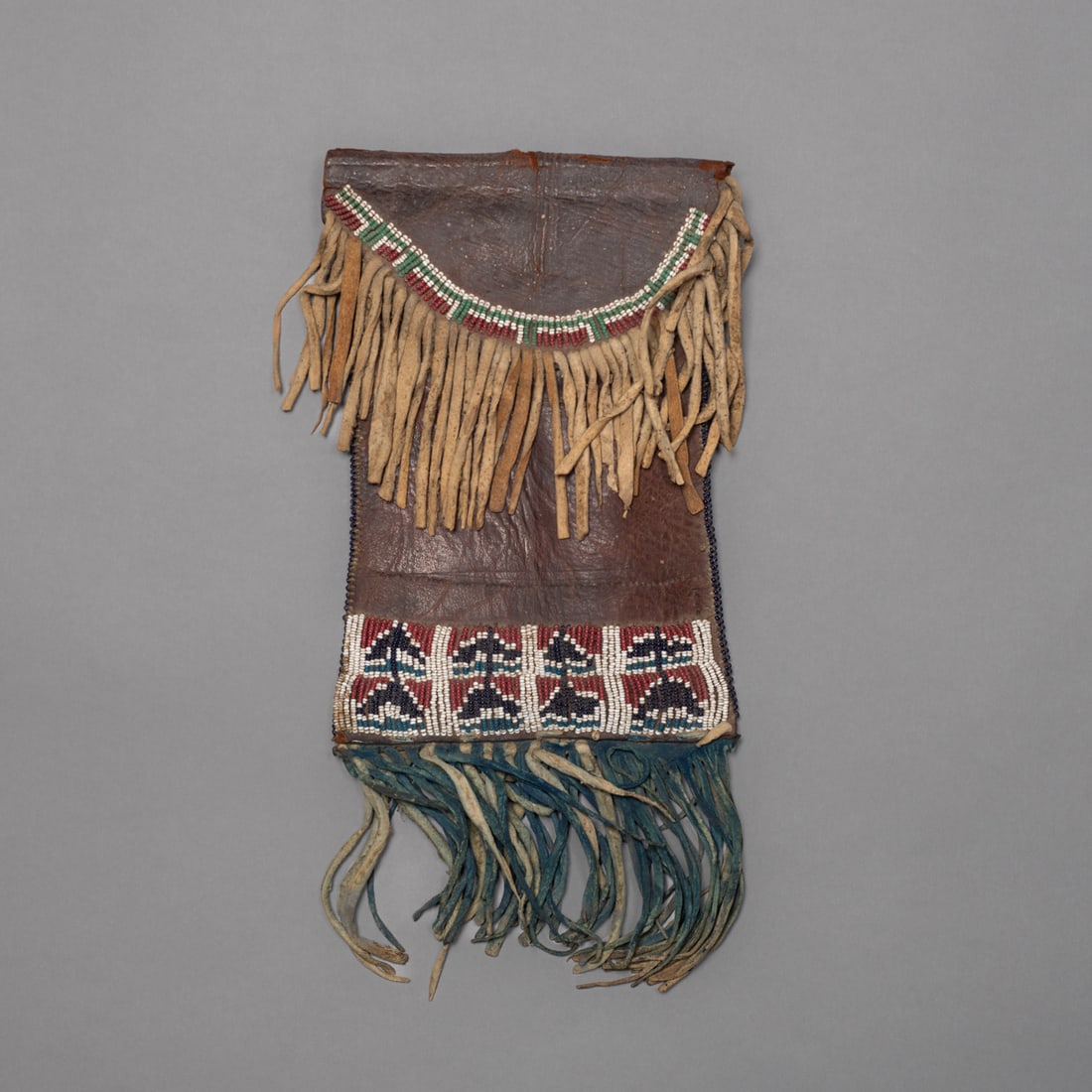 An early Kiowa dispatch case: Southern plains, circa 1860s early 1870s, native beaded on oak dyed commercial saddle hide with native tanned fringe. The beaded strip formed with four rectangular panels of navy, blue and red on a