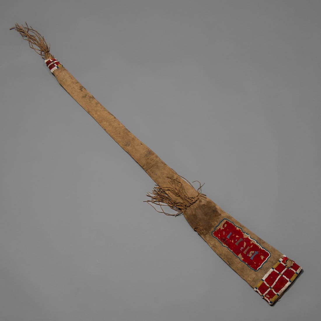 A Northern Plains pony beaded gun case: The elongated tanned hide gun case featuring classic Northern Plains design elements, with sinew-sewn pony beadwork panels at each end and a central red trade cloth field bordered in blue, greasy