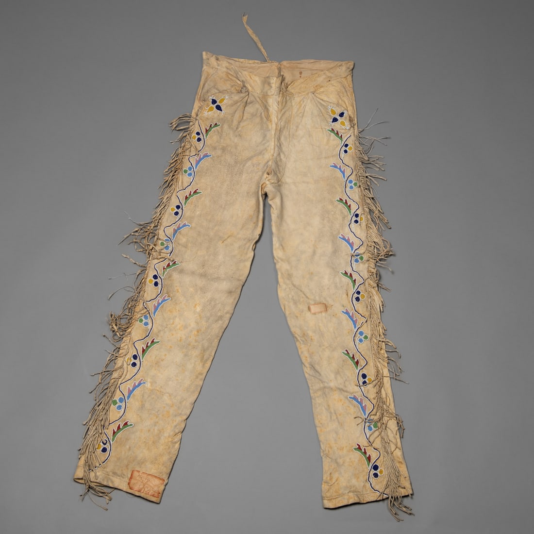 A pair of Northern Plains beaded scout pants: 19th century, decorated along the outer seams with classic floral beadwork in blue, green, yellow, red, and white on a light hide ground. Long fringe runs the full length of each leg, with remnants