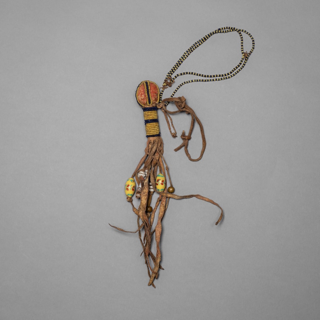 A Crow or Blackfoot personal hair adornment: Late 19th century, composed of hide, beadwork, and trade embellishments. The upper section features a red cloth-wrapped core encircled by yellow and navy seed beads in banded rows. From it extends a