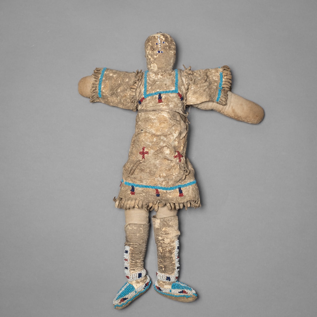 A Northern Plains hide doll: A Lakota doll constructed of stuffed hide, dressed in a beaded dress and leggings. The beadwork executed in glass seed beads of white, blue, red, and dark navy forms geometric motifs. The doll also