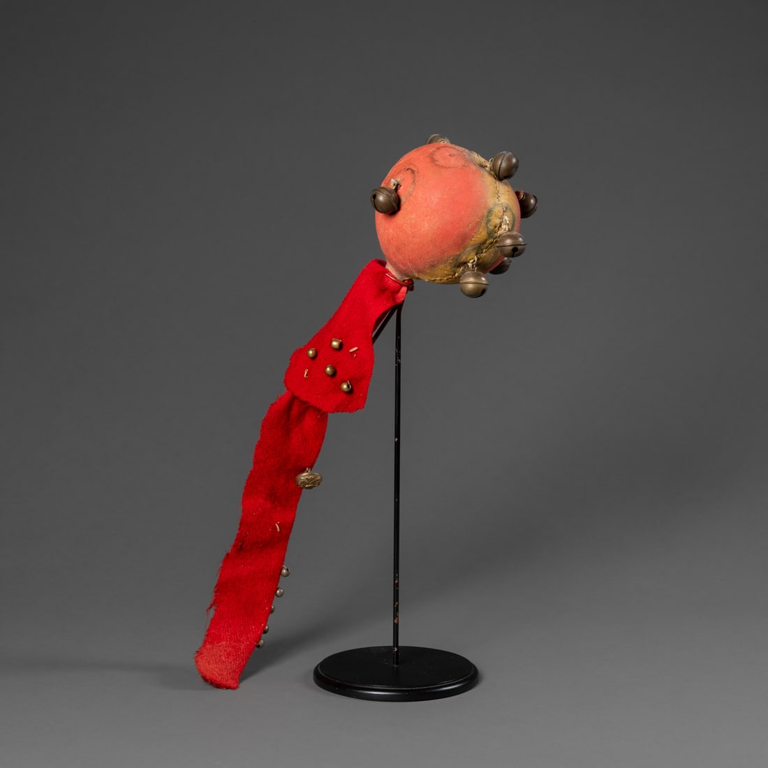 A Crow globular rattle: C. 1860-80, the head wrapped with painted hide, with a number of brass bells attached, the wood handle wrapped in a long piece of red trade cloth, with brass beads and a military button attached,