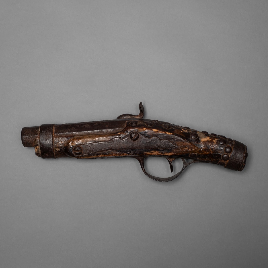 A Plains Indian blanket gun: Short and thick wooden and metal gun, darkened with age and covered in iron fittings and decorated with tacks. The metal has a deep brown patina from age, and the wood has a rough, hand-carved