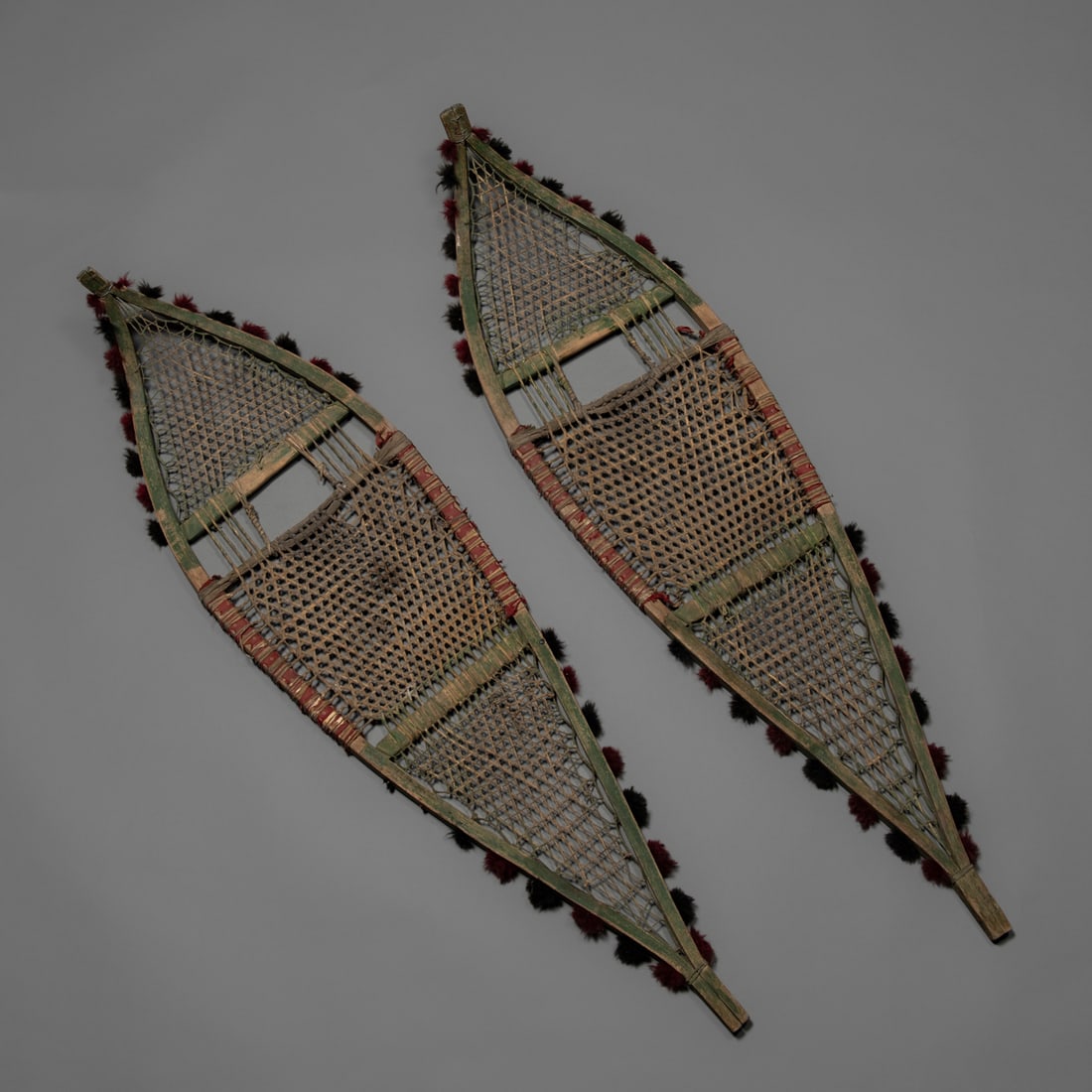 A pair of Western Great Lakes snow shoes: Ojibwa/Chippewa/Winnebago, or Sauk Fox, intricately wrapped with leather cordage, the wooden supports adorned with wool poms on each side, remnants of old red trade cloth around the middle sections.
