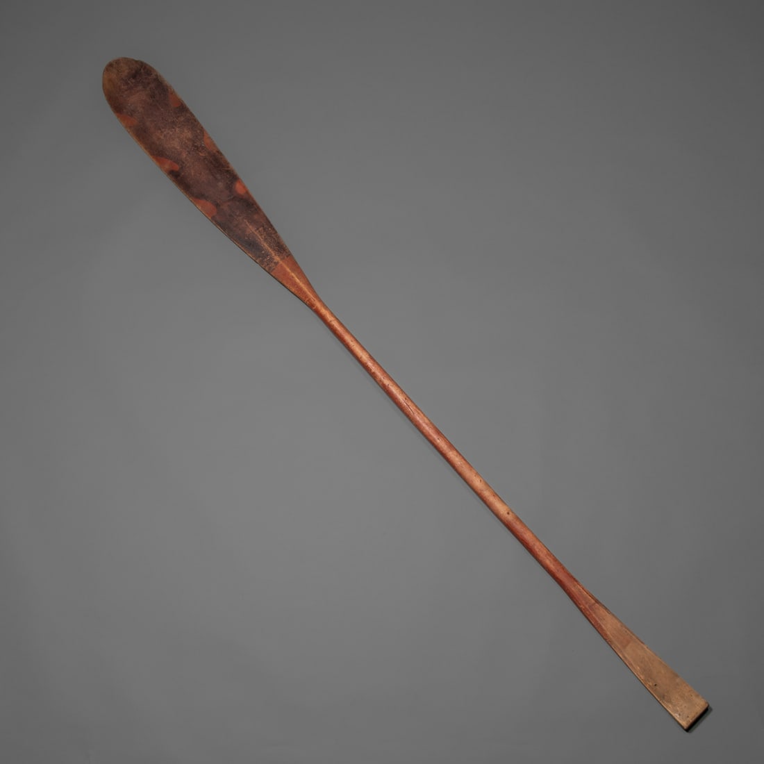 A Northeast woodland canoe paddle: Hand-carved wooden canoe paddle featuring an elongated blade form with remnants of original red and black pigment. The surface shows a warm, softened patina, and the tapered handle retains signs of