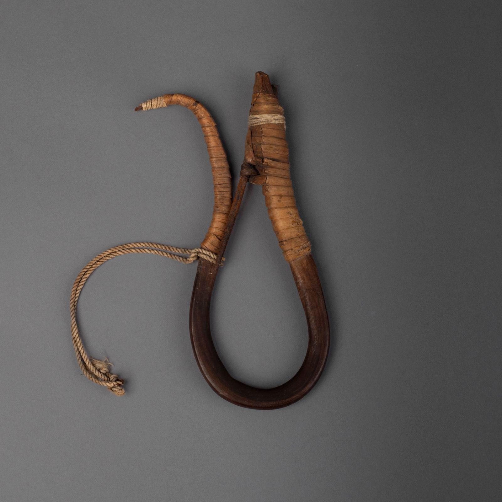 A Northwest Coast bentwood halibut hook: Haida/Tlingit, 19th century, the bent wood with cedar bark wrapping, with an iron barb and cord, dark brown patina to the wood. Lg. 6 1/2 in.