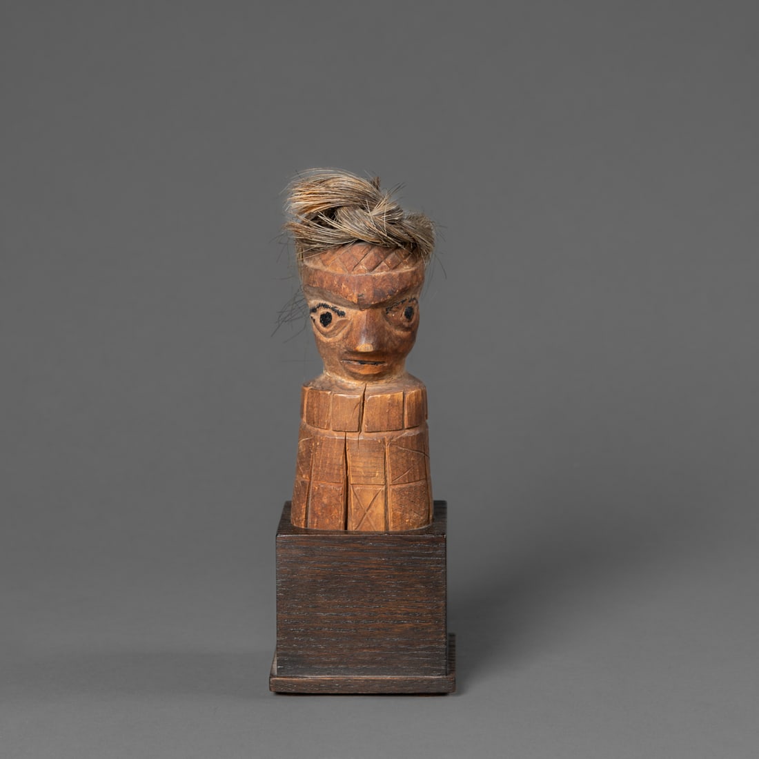 An Early Northwest Coast wood carved figure: Nootka, Nuu-chah-nulth, early 19th century, the wood head sits on a raised columnar base with cross-hatched incising, the head with large eyes painted black, a sharp nose and animal hair on the head,
