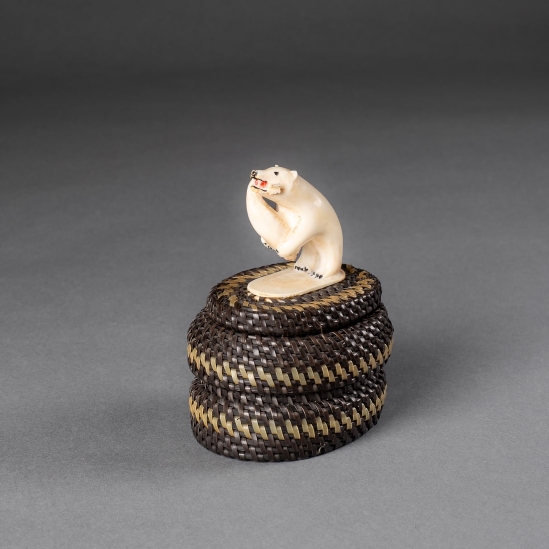 A contemporary Point Hope Baleen basket: L. Koonok Sr. A baleen basket of ovular form, the lid surmounted by a dynamic carving of a polar bear grasping a fish in his mouth, finely detailed with painted eyes and red pigment at the mouth.