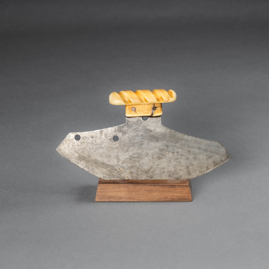 A Bering Sea Eskimo woman’s knife, ulu: Crescent-form iron blade fitted with a carved handle featuring a twisted grip design. The blade features holes for attachment points. Comes in a custom wooden stand for display. Wd. 7 ½, ht. 4