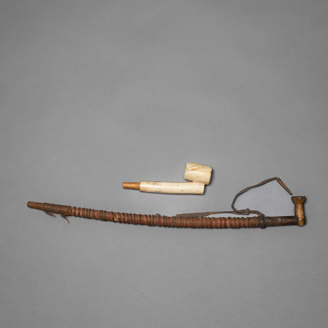 Two Eskimo pipes: Alaska, late 19th century, constructed with a slender wooden stem tightly wrapped in sinew, fitted with a small carved bowl at the upper end. Retains attached hide thong and shows deep patina from