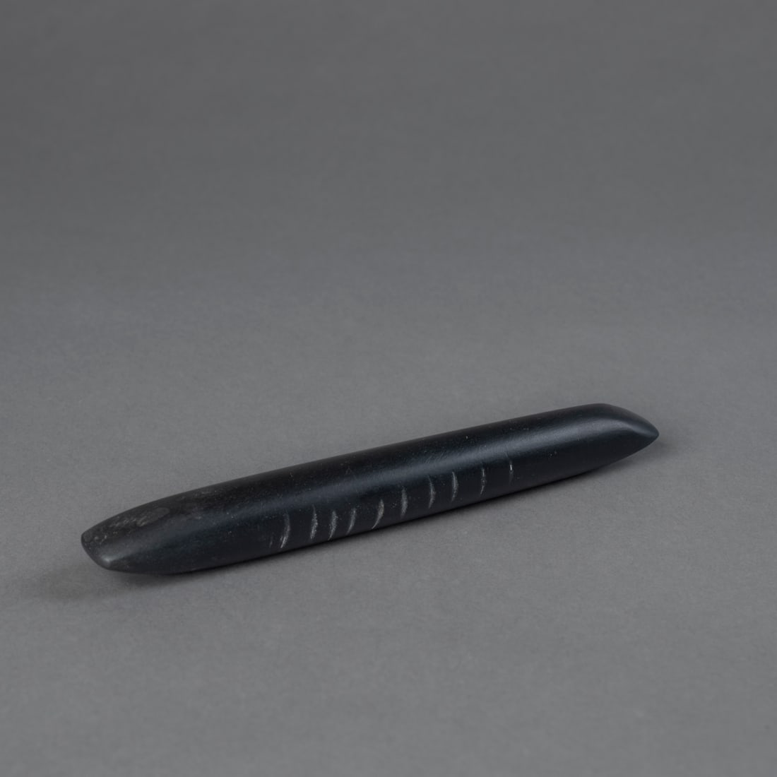 A Pueblo black stone tool: Polished black stone tool, with tapering ends and grooves along the midsection, exhibiting a smooth surface. Lg. 7 ¼ in.