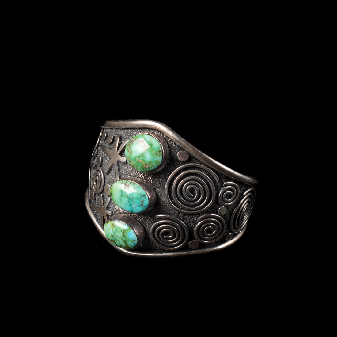 An Alex Sanchez silver and turquoise cuff bracelet: The heavy Sterling silver bracelet with petroglyph designs and three turquoise settings in raised bezels, signed "Alex Sanchez," and marked "Sterling." Total interior circumference (including gap) 6 3