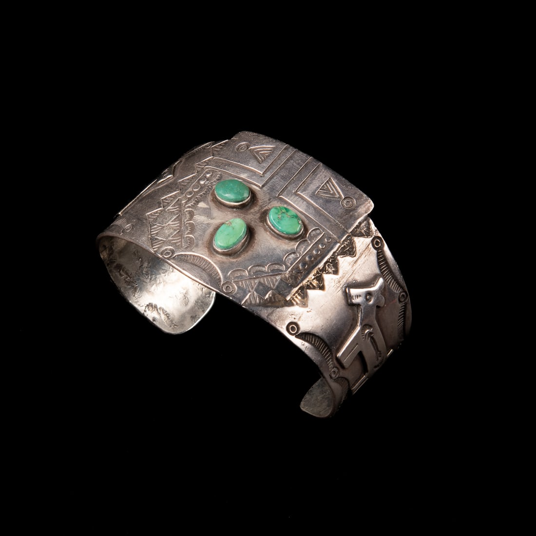 A Fred Harvey silver and turquoise Kachina Mask bracelet: The silver cuff with stamp work two overlay dogs and a plaque of a Kachina mask with three turquoise settings. Total interior circumference (including gap) 6 5/8, wd. 1 1/2, gap 1 1/8 in. Gross weight