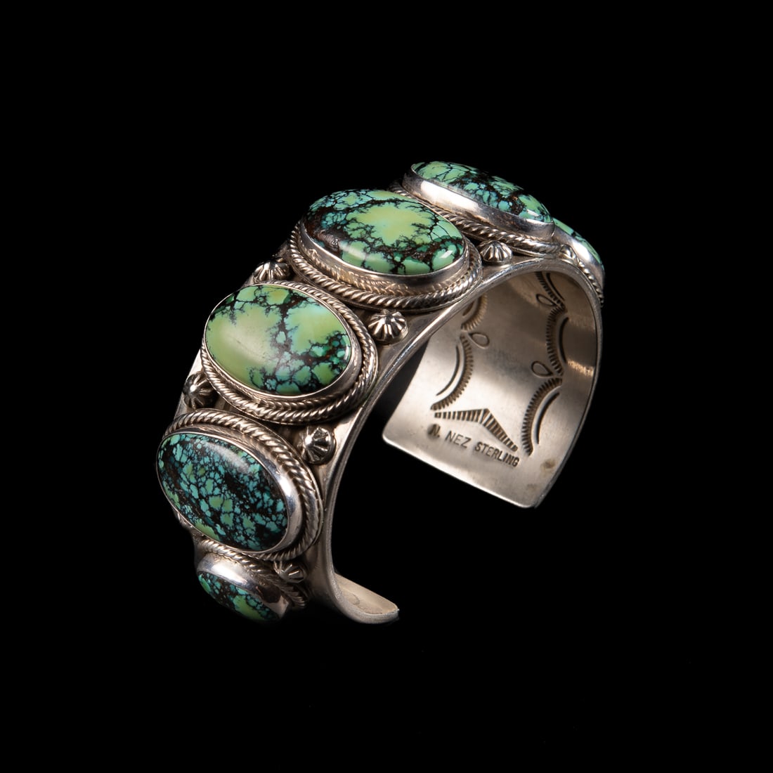 A Ned Nez silver and turquoise cuff bracelet: The wide Sterling silver cuff with a row of seven graduated Spiderweb turquoise settings with rope and star additions around the stones, the inside decorated with stamp work, signed, "N. Nez," and mar