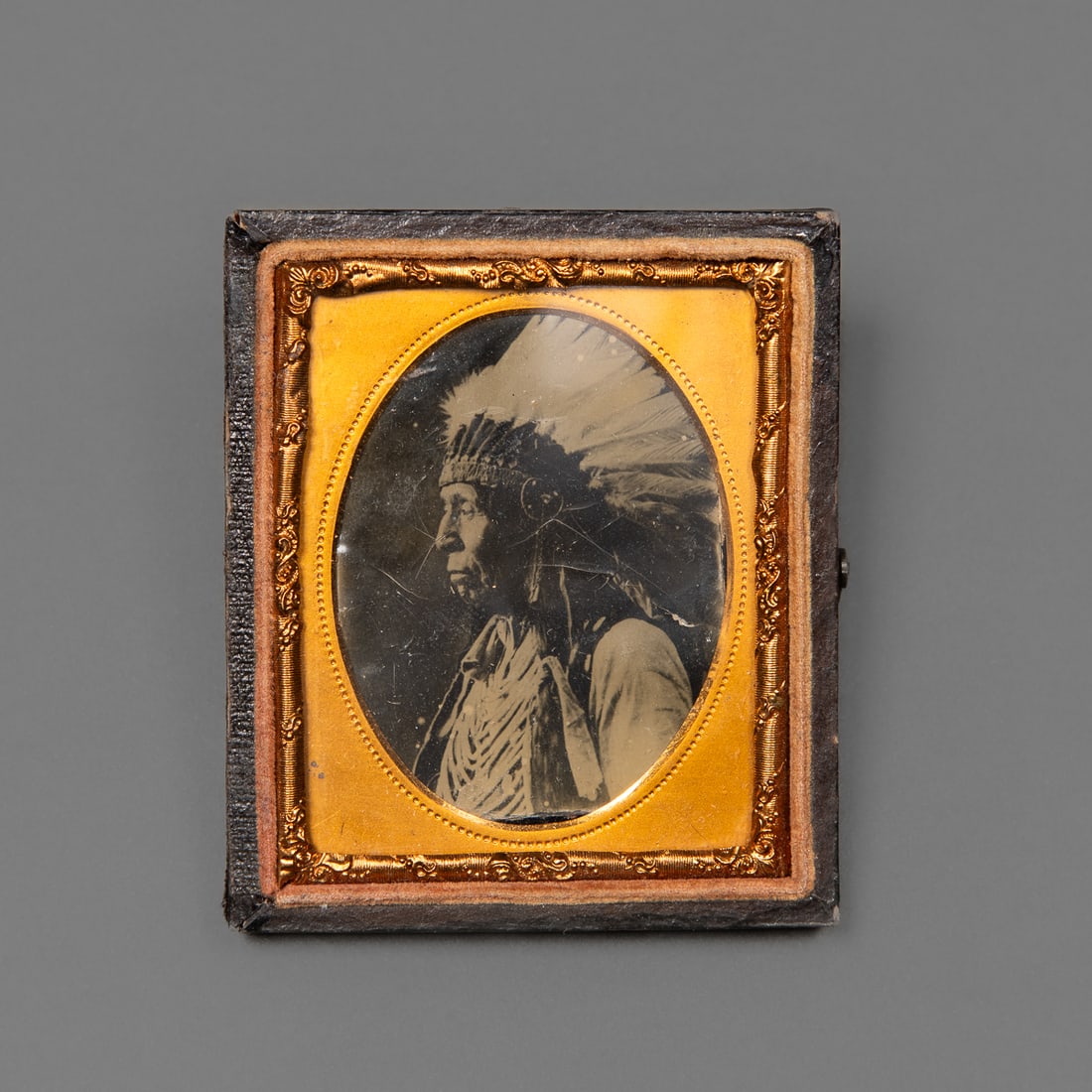 A Ferro/Tintype photograph of an Indian chief: A 19th-century tintype or ferrotype portrait depicting a Native man wearing a feathered headdress and traditional attire, housed in its original gilded brass mat and pressed leather case. Ht. 2 7/8, w