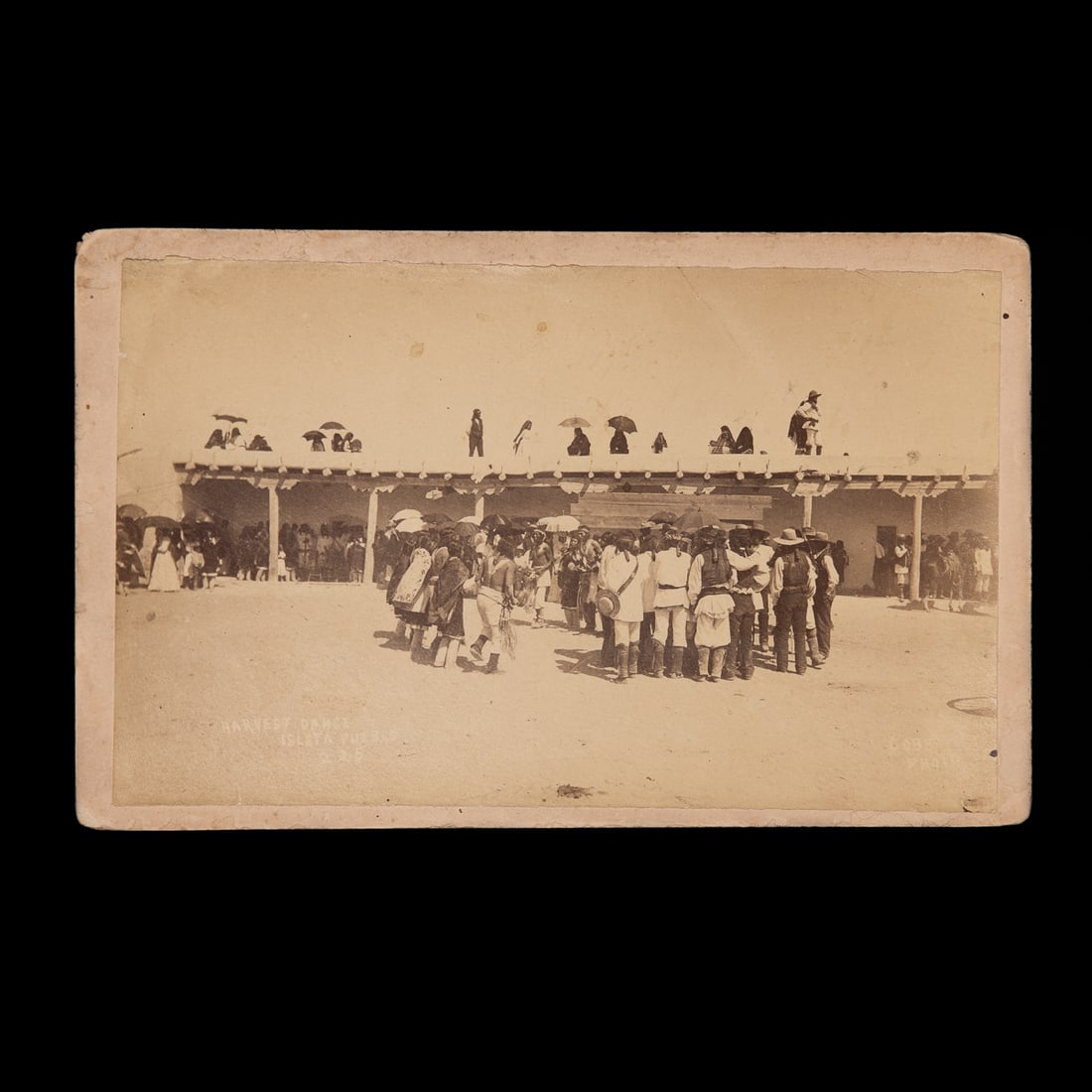 A cabinet card depicting a large group of Pueblo Indians: H.W. Wyman Isleta Pueblo Harvest Dance albument print, large cabinet card, Santa Fe, depicting a large group of Pueblo Indians gathered in the plaza of Isleta Pueblo for their annual Harvest Dance. Ht