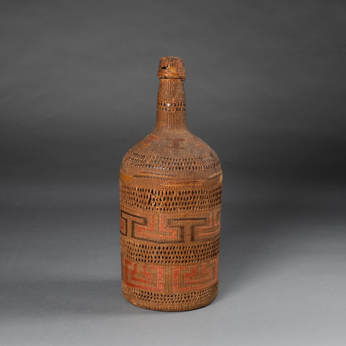 A large Tlingit basketry bottle: Tlingit, c. 1900, finely woven basketry bottle, constructed in natural, dyed spruce root, and maidenhair fern. The cylindrical form rises to a long, narrow neck, capped with a fitted woven stopper.