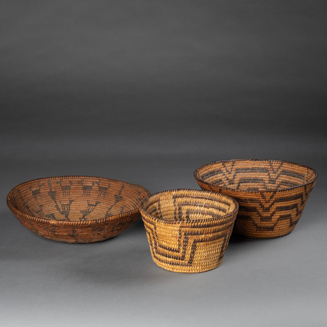 Three Southwest baskets: Each finely coiled with distinct geometric and radiating designs in natural and dyed vegetal fibers. The smaller two baskets featuring meander motifs, the larger with animals depicted around the