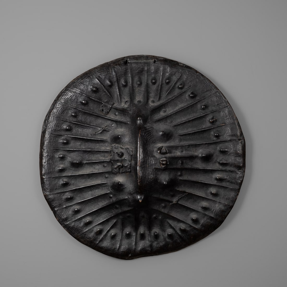 An Ethiopian Arussi hide shield: Of asymmetrical circular form, the thick hide with embossed raised bumps radiating from a central raised vertical spine, pierced through at the center for the attachment of a leather handle, varied