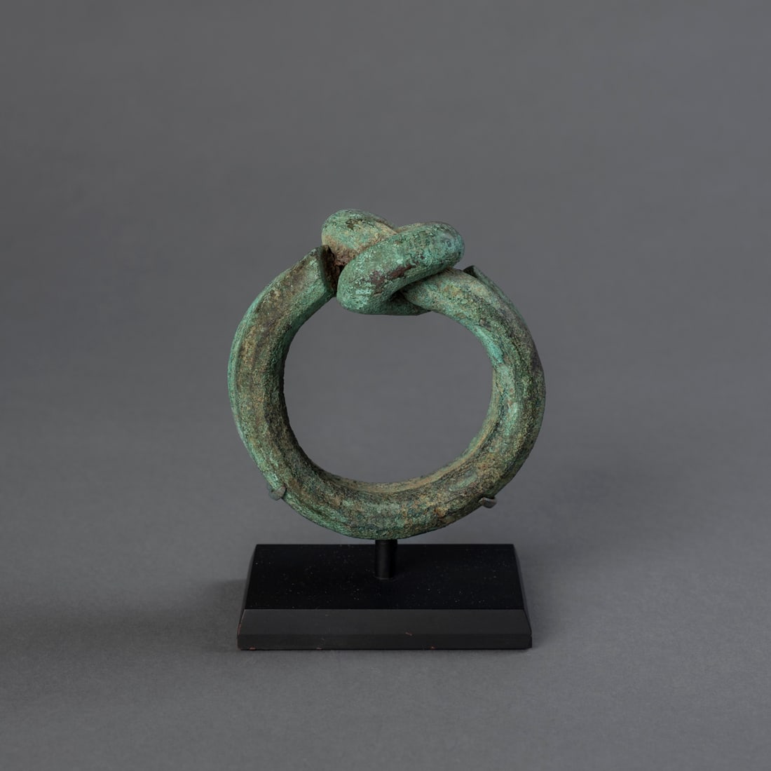 A Nigerian copper currency knot bracelet: Cast in heavy copper, worked into a broad circular band with the upper section formed into an interlocking knot, the surface with a deep green patina. Mounted on a metal stand. Ht. 5 with stand, dia.