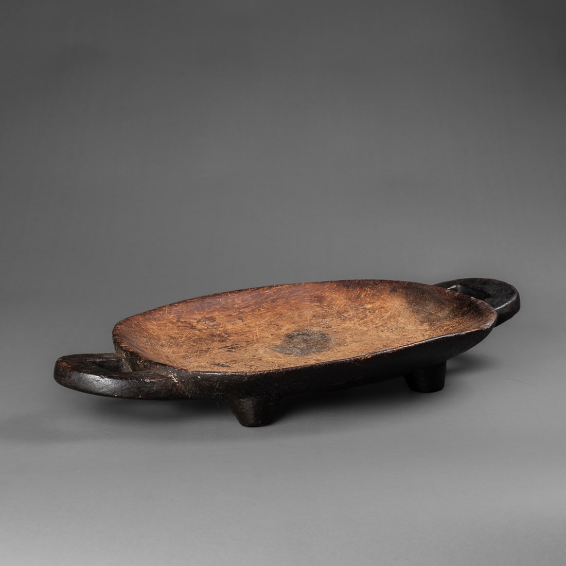 A Zulu wood food platter: Zulu, 19th century, the shallow dish with four short legs, the underside decorated with two raised triangles, each end with large extended handle, the underside with encrusted black patina. Ht. 2