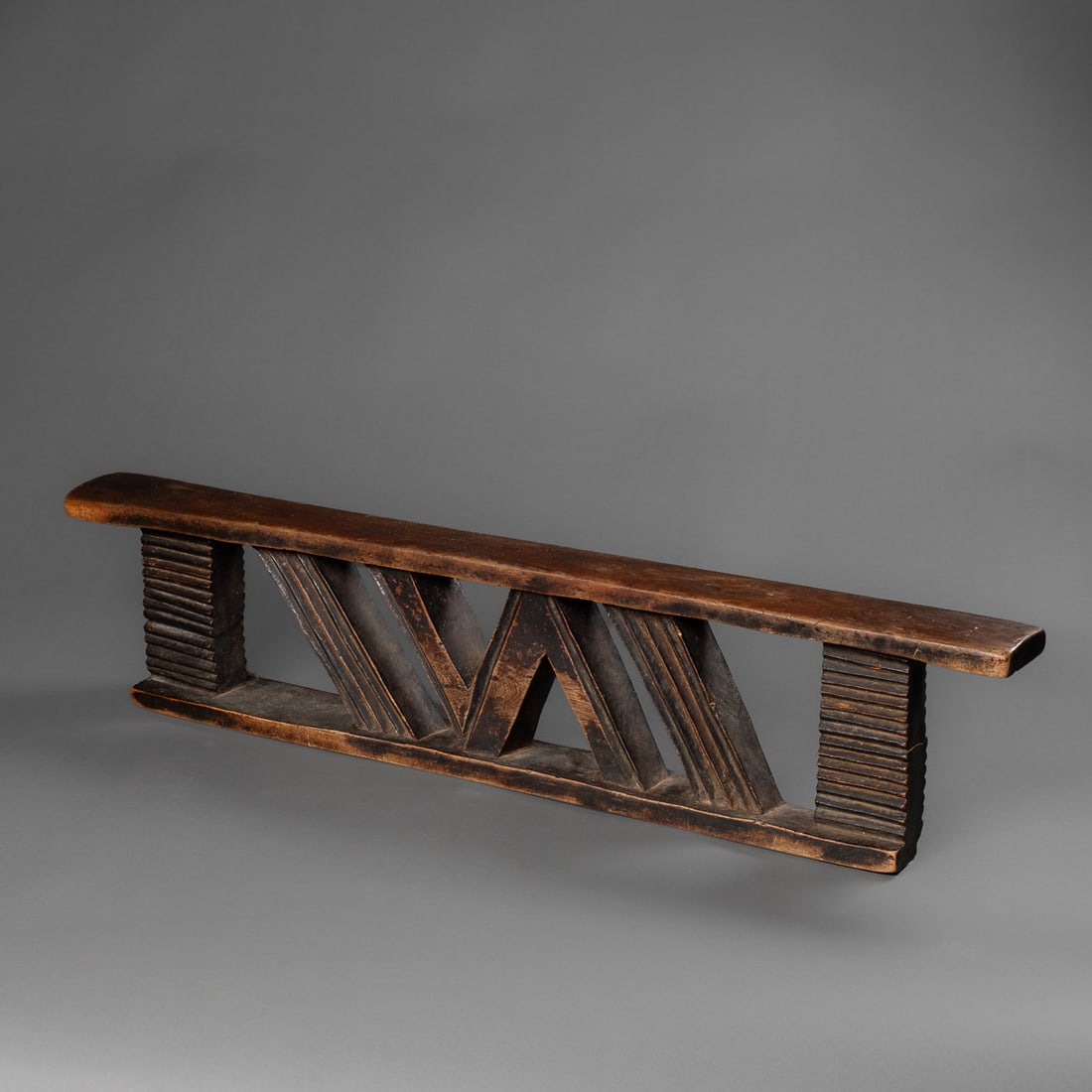 A South African Neck rest: Zulu, 19th century, the long narrow wood base supports two rectangular columns with deep incised grooves the center with two slanting grooved columns and a central “N” support. Smooth dark