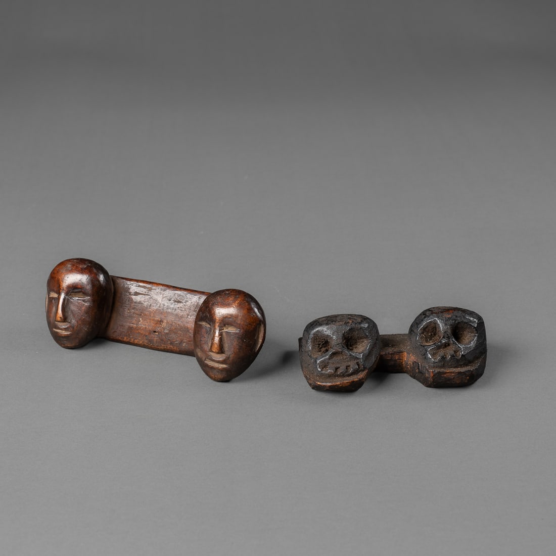 Two Naga double heads: Konyak, the hand carved wooden double heads featuring two faces with one ear each, the other double head carving features skull imagery with three-hole attachment points. Both feature a rich dark
