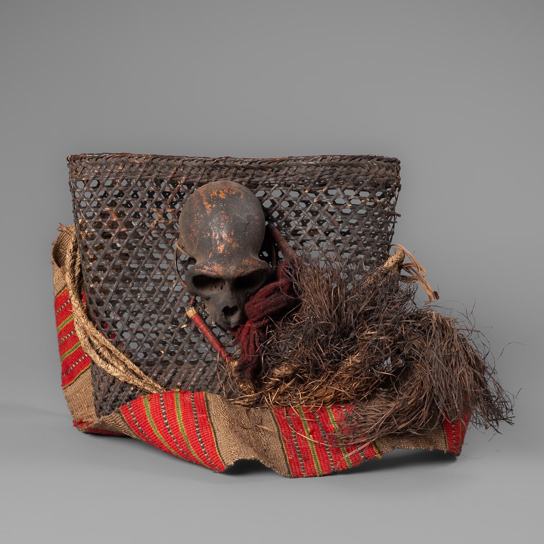 A small Naga headhunter basket: Konyak, the woven basket with a thick woven baldric, decorated with red, yellow, green, and brown pigment that branches into three separate ties. Attached to the bag are six beaded, cloth wrapped,