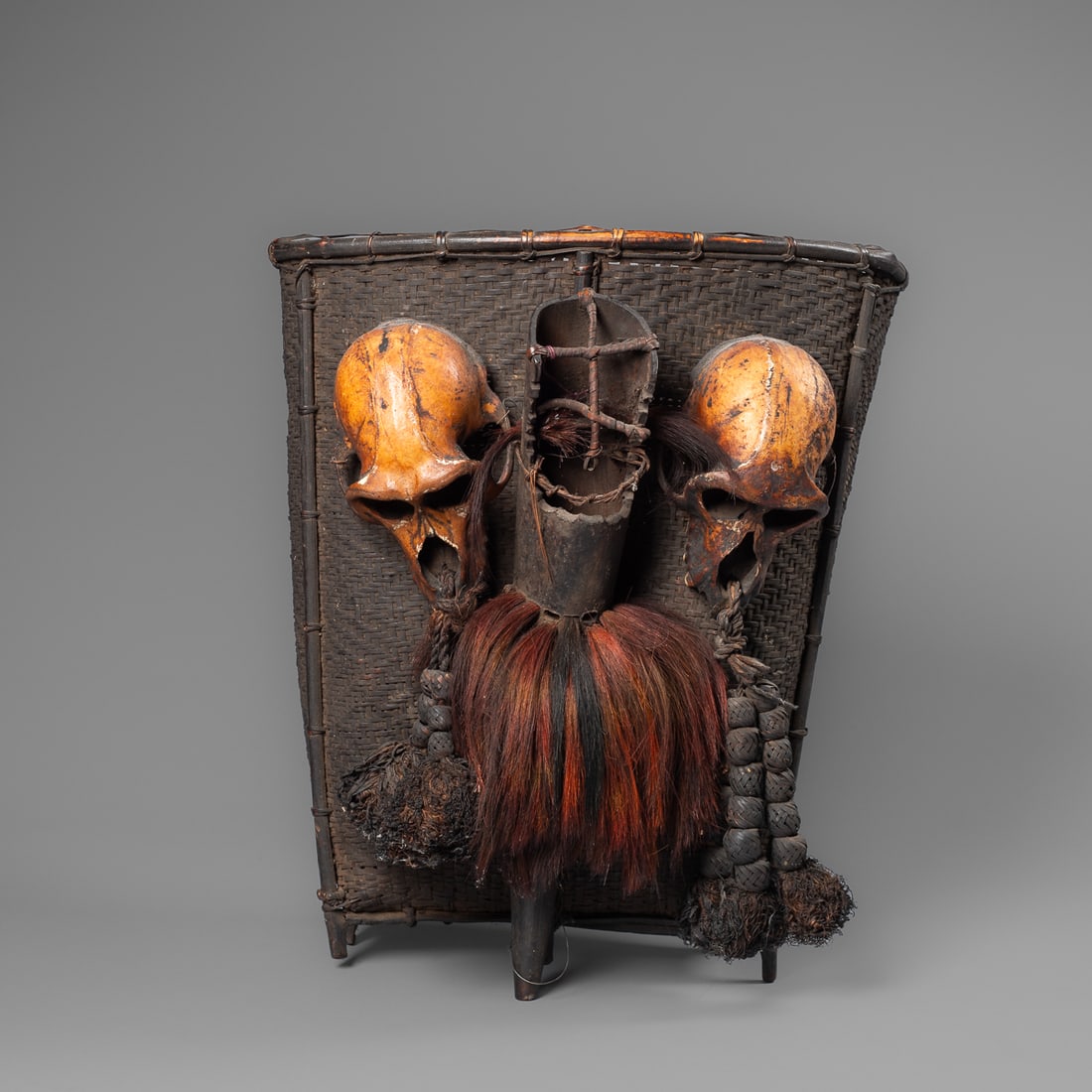 A large Naga headhunter basket: Konyak, the woven basket with five bottom supports. Attached to the bag are two monkey skulls with a dark orange patina. The bottom of the skulls has woven dangles attached through the nose slit. The