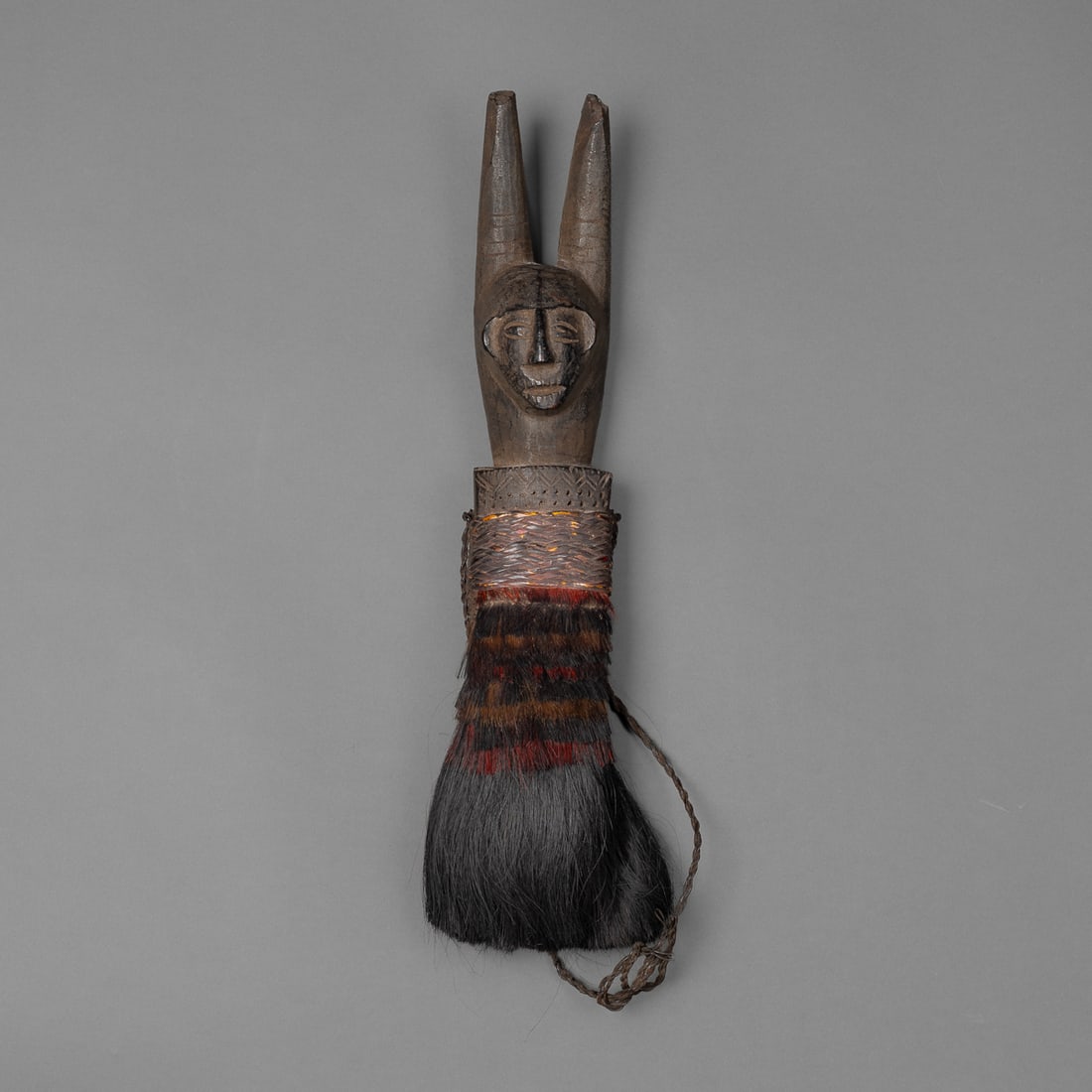 A Naga hip ornament with wood face: The carved wooden figure with two forked horns protruding from its head, with detailed face, teeth and open mouth. The base with incised decorative lines and dotting, wrapped with woven basketry and