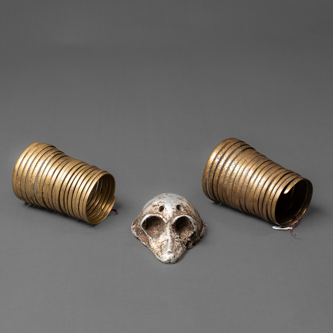 Two brass wire Naga gauntlets & aluminum skull: The two brass wire gauntlets, made by shaping around a wooden frame, are incised with geometric hatching patterns. The brass coil contains a shiny patina. The aluminum skull is with incising on the